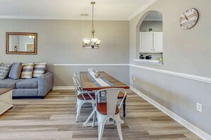 Ein Essbereich mit Holztisch, weißen Stühlen, einer Chandelier und einem grauen Sofa mit gemusterten Kissen.