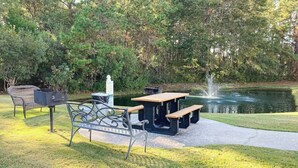 Un espace extérieur aménagé avec un barbecue, une table et des bancs, situé près d’un étang.