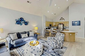 Ein modernes Wohnzimmer mit einer Couch, einem Hocker und einem gemusterten Sessel. Im Hintergrund ist eine Küche mit Holzausstattung und ein Essbereich mit Stühlen erkennbar.