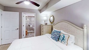 Quarto com cama, ventoinha no teto, mesa de cabeceira, espelho e porta para um banheiro.