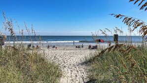 Uma praia com céu claro, um caminho de areia que leva à água, e algumas pessoas na praia.