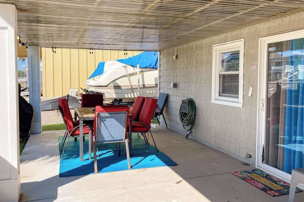 Un patio cubierto con una mesa y sillas, un barco al fondo, y una puerta que da al interior.
