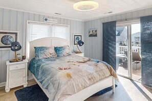Ein Schlafzimmer mit einem Bett, einem Nachttisch, einer Lampe, einem Fenster mit Jalousien und Blick nach draußen.