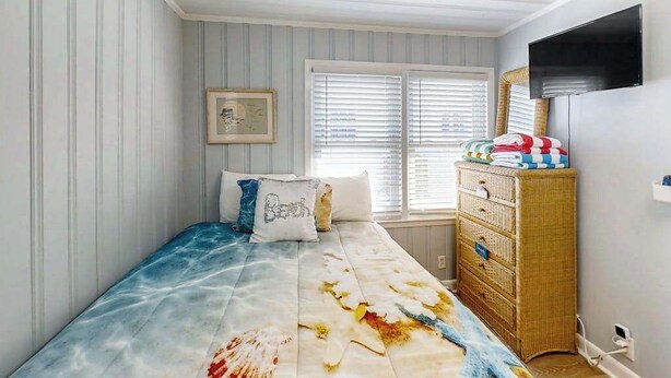Een slaapkamer met een bed, een dressoir, een televisie en een raam met jaloezieën.