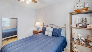 A bedroom with a bed, a nightstand, a lamp, a ceiling fan, and a shelf with model ships. - 3rd Avenue Oasis Retreat (North Myrtle Beach)