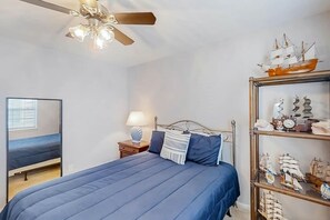 A bedroom with a bed, a nightstand, a lamp, a ceiling fan, and a shelf with model ships. - 3rd Avenue Oasis Retreat (North Myrtle Beach)