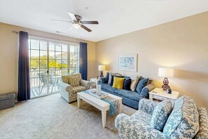 Ein Wohnzimmer mit einer Couch, einem Sessel, einem Couchtisch und einem Deckenventilator. Ein Fenster mit Vorhängen bietet einen Blick ins Freie.