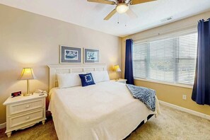 A hotel room with a bed, nightstand, lamp, ceiling fan, window with blinds, and wall art. - Barefoot Beach Retreat (North Myrtle Beach)