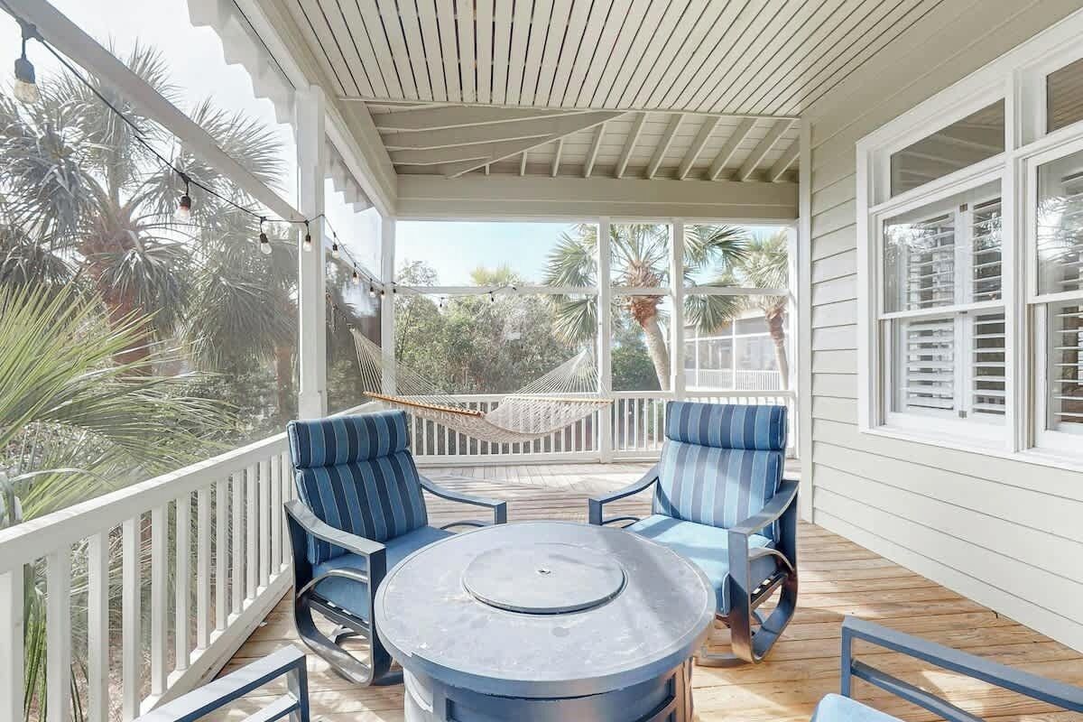 A covered outdoor seating area with a round table and blue chairs, surrounded by a white railing and palm trees.