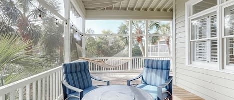A covered outdoor seating area with a round table and blue chairs, surrounded by a white railing and palm trees.