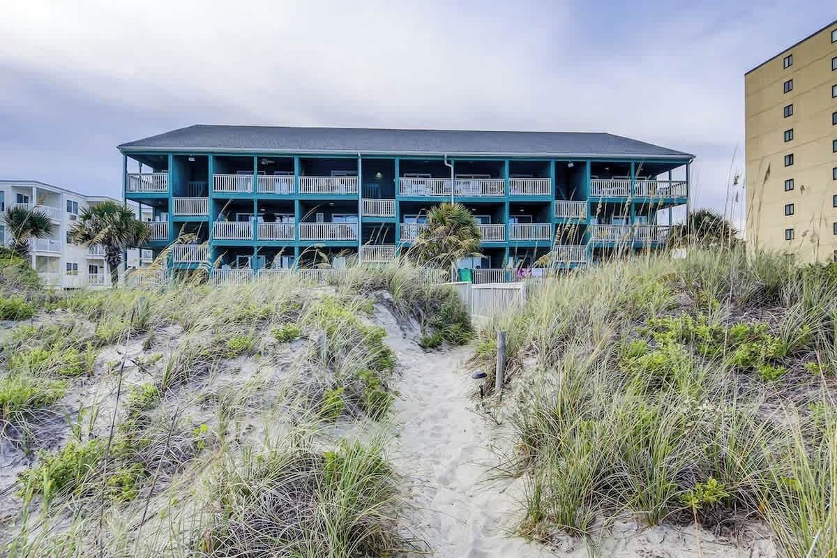 A multi-story building with balconies, a sandy path leading to it, and a beach in the background.