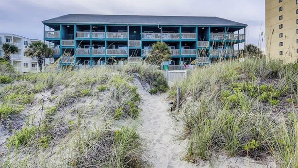 Un edifici de diverses plantes amb balcons, un camí de sorra que hi convergeix i una platja al fons.