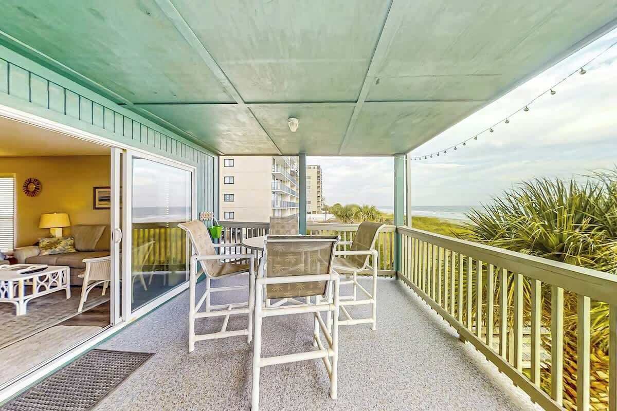 A covered outdoor area with a table and chairs, a view of the ocean, and a building in the background.