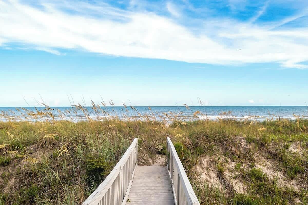 A wooden boardwalk leading to a beach with grassy dunes and a clear sky.