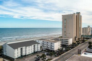 A coastal hotel with a parking lot, a beach view, and a tall building in the background. - Windcrest Villas 201 (North Myrtle Beach)