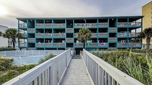 Un edifici de diversos pisos amb balcons, un camí de fusta que condueix a l'entrada i una palmera al primer terme.