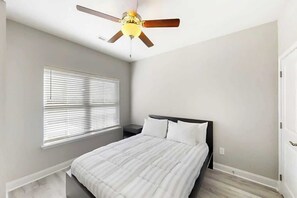 A bedroom with a bed, a ceiling fan, a window with blinds, and a door. - Cocora on Church Street - A (Myrtle Beach)