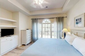 A bedroom with a bed, a television on a dresser, a window with curtains, and a lamp on a bedside table. - Cypress Bend Unit 432 (North Myrtle Beach)