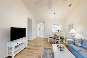 Uma sala de estar com uma televisão de tela plana sobre um suporte branco, um sofá azul, uma mesa de centro e uma área de jantar com mesa e cadeiras.