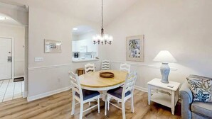 A dining area with a round table, white chairs, a chandelier, a side table with a lamp, and framed artwork on the walls. - Cypress Bend Unit 432 (North Myrtle Beach)