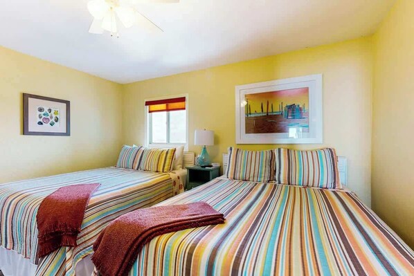 Habitación de hotel con dos camas, un ventilador de techo, una ventana con persiana roja y blanca, y un cuadro en la pared.