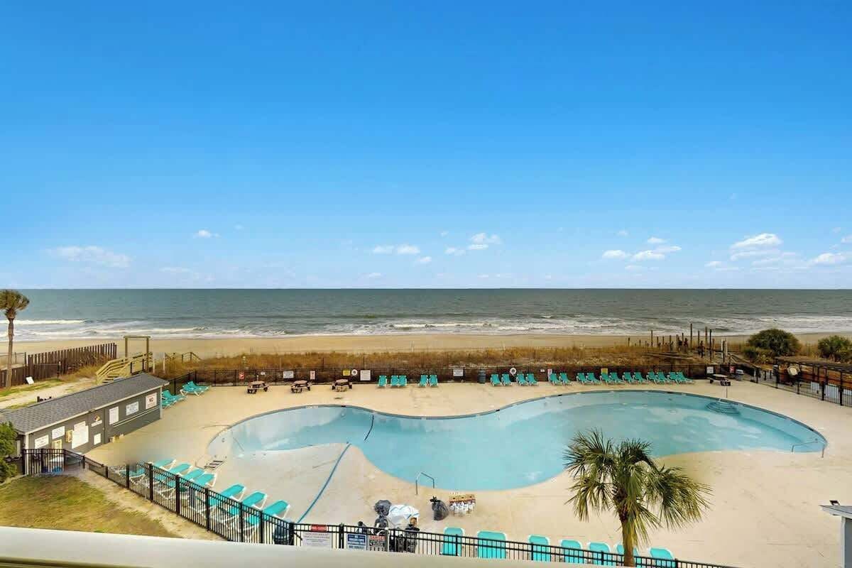 A swimming pool with a curved design, surrounded by beach chairs and a palm tree, with a beach and ocean in the background.