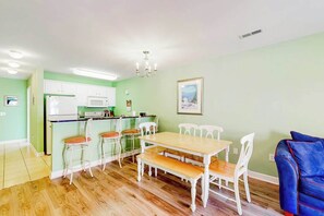 A dining area with a wooden table and white chairs, a kitchen with white cabinets, and a blue sofa. - The Singing Gull Retreat (Myrtle Beach)