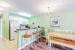A kitchen with white cabinets, a refrigerator, a microwave, and a dining area with a wooden table and white chairs. - The Singing Gull Retreat (Myrtle Beach)
