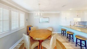 A dining area with a round wooden table surrounded by wicker chairs, a kitchen with white cabinets, and a bar area with stools. - *Hook Wine and Sinker (Surfside Beach)