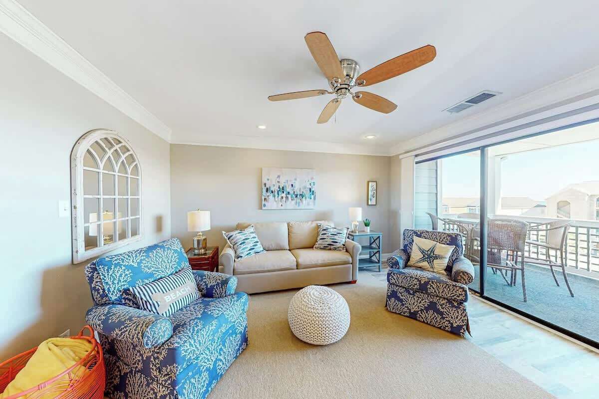 A living room with a ceiling fan, a sofa set, a round ottoman, and a view of the outdoors through large windows.