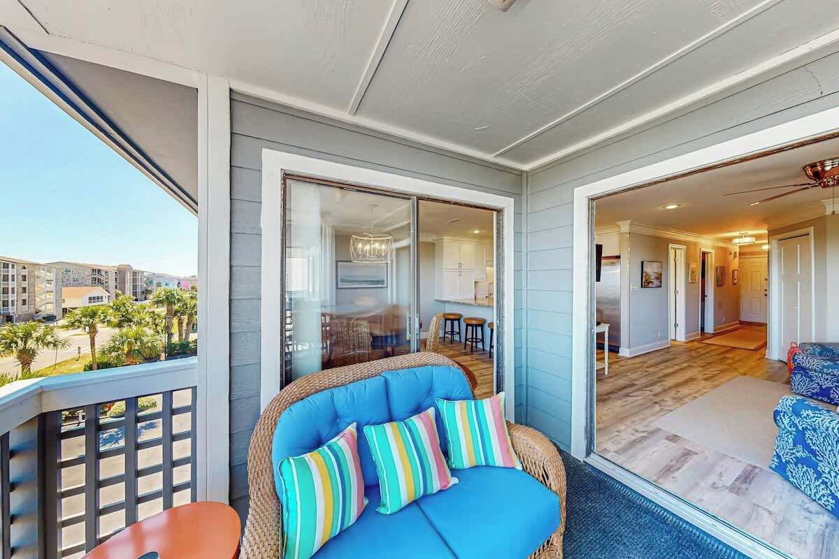 A balcony with a wicker sofa, colorful cushions, and a view of a residential area.