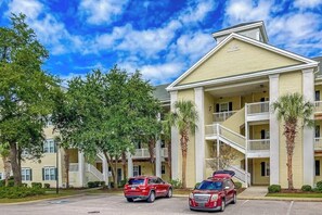 A multi-story hotel with a central courtyard, balconies, and a parking area with two cars. - Ocean Keyes 3722 The Gleaming Starfish (North Myrtle Beach)