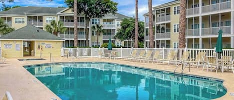 A swimming pool with a white fence, surrounded by multi-story buildings with balconies, under a clear sky.
