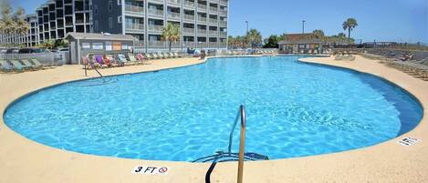 Una piscina con un indicador de profundidad de 3 pies, rodeada por una playa de arena y un edificio de varios pisos al fondo.