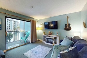 Un salon avec un canapé, un téléviseur, une guitare et un balcon équipé de chaises.