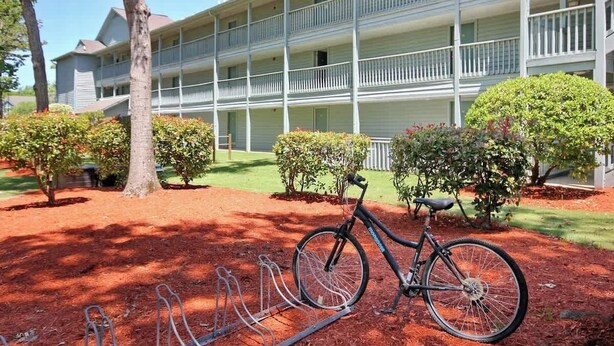 A multi-story building with a balcony on each floor, a bicycle parked in front, and a well-maintained garden with trimmed bushes. - The Golden Sailboat B-139 (Myrtle Beach)