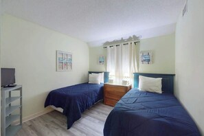A bedroom with two single beds, a bedside table, a TV stand, and a window with curtains. - The Golden Sailboat B-139 (Myrtle Beach)