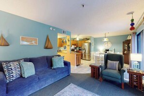 A living room with a sofa, armchair, and a small table. There is a kitchen in the background with a refrigerator and cabinets. - The Golden Sailboat B-139 (Myrtle Beach)