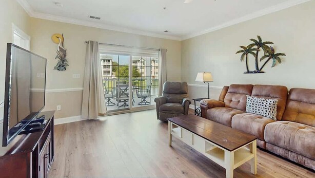 A living room with a flat-screen TV, a sofa, a coffee table, a ceiling fan, and a balcony with outdoor furniture. - Turtle Cove Unit #204 (Myrtle Beach)