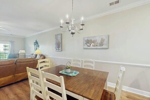 A dining area with a wooden table, white chairs, a chandelier, and artwork on the walls. - Turtle Cove Unit #204 (Myrtle Beach)