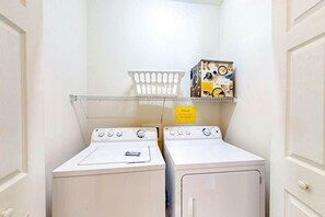 A laundry room with two white washing machines, a metal drying rack, and a shelf with a box of laundry detergent. - Turtle Cove Unit #204 (Myrtle Beach)