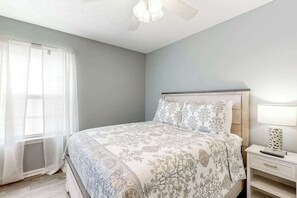 A bedroom with a bed, a nightstand, a lamp, and a window with sheer curtains. - Sunny Shores Retreat (Myrtle Beach)