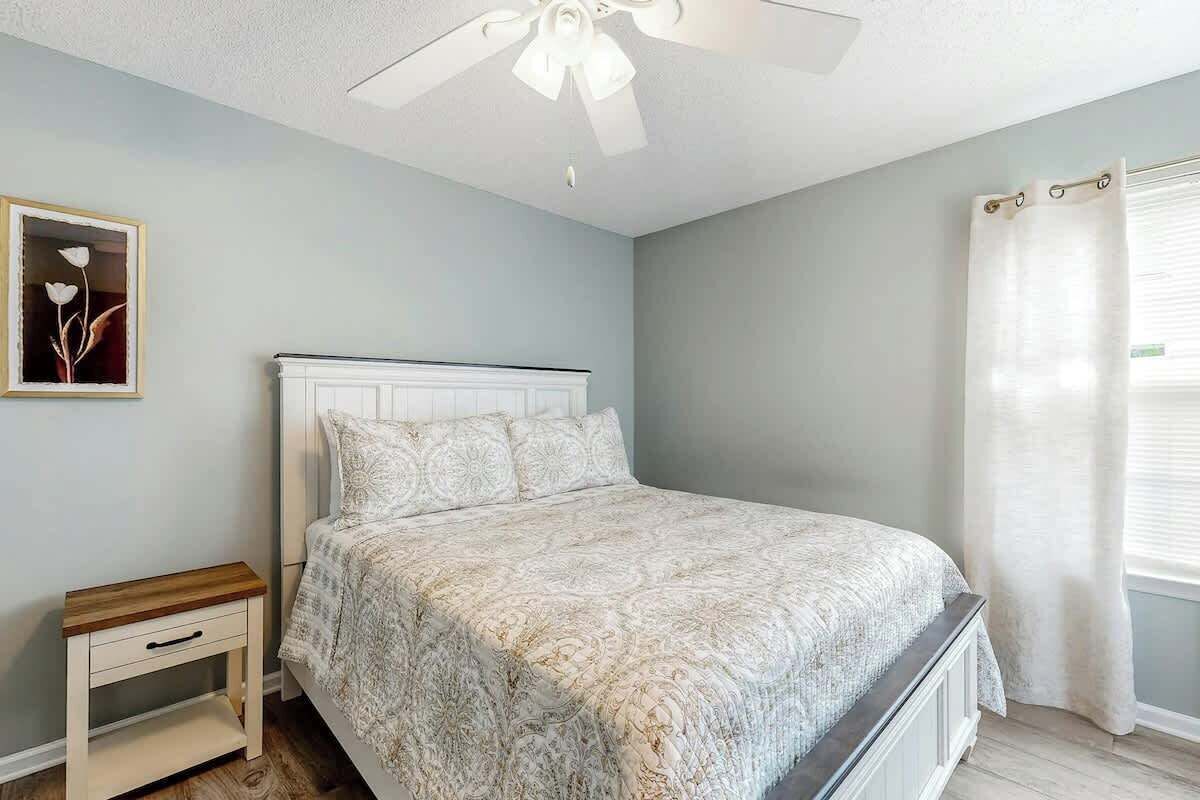 A bedroom with a bed, a nightstand, a ceiling fan, a window with curtains, and a wall-mounted picture.