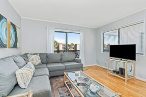A living room with a grey sofa, a glass coffee table, a flat-screen TV on a stand, and a large window with blinds. - Inlet Point Villas 10-D (North Myrtle Beach)