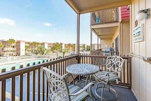 A balcony with a round table and two chairs, overlooking a swimming pool and a street with palm trees. - Inlet Point Villas 10-D (North Myrtle Beach)