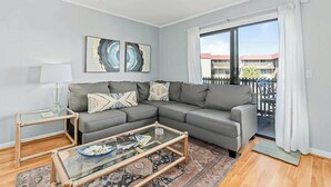 A modern living room with a grey sectional sofa, a glass coffee table, and a view of a balcony with a railing. - Inlet Point Villas 10-D (North Myrtle Beach)