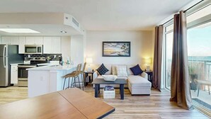 En moderne stue med sofa, sofabord og spisebord. Der er et køkkenområde med hårde hvidevarer og et stort vindue med udsigt.