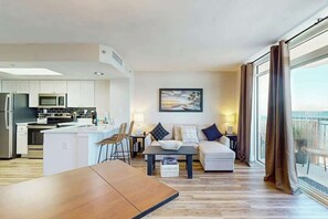 A modern living room with a sofa, coffee table, and dining table. There is a kitchen area with appliances and a large window offering a view. - Sandy Daydreams, Unit PH3 (Myrtle Beach)