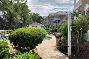 A pool area with lounge chairs, a small building, and a well-maintained garden. - Gardens at Cypress Bay 14-204 (Little River)