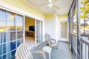 Ein Balkon mit Deckenventilator, eine Glastür, die zu einem Raum mit Fernseher führt, und zwei Adirondack-Stühle.